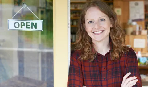 A young woman entrepreneur smiles and leans against the door of her shop which displays an "open" sign